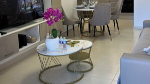 Una sala de estar moderna con un televisor de pantalla plana, un comedor con mesa para dos personas, y una cocina al fondo.