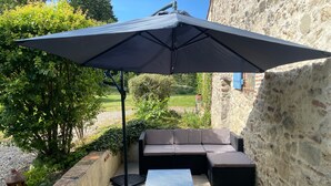 An outdoor patio with a black umbrella, a sofa, and a table. - La Maison du Jardin – Country Estate with Large Pool and Wi-Fi (Penne-d'Agenais)