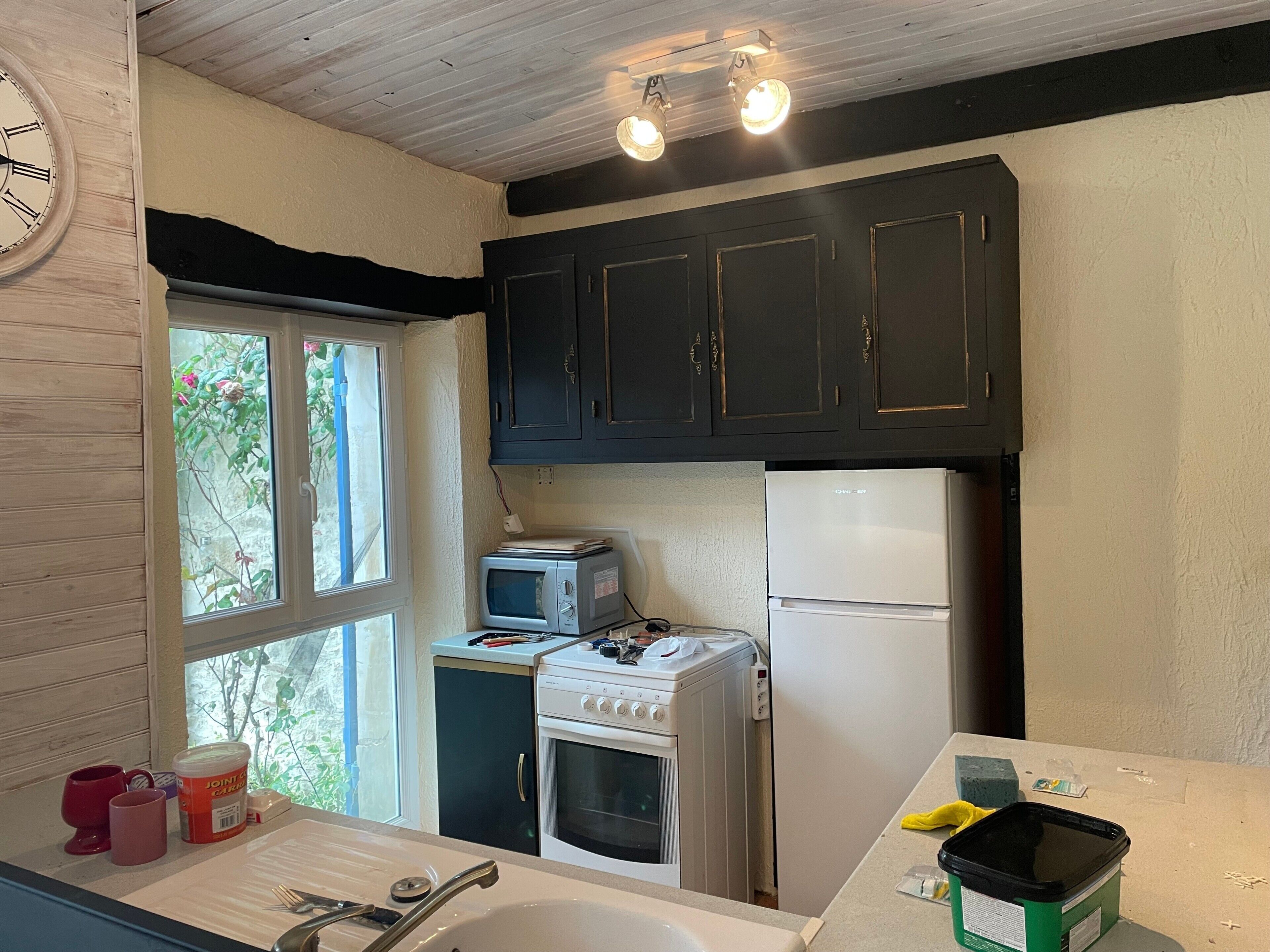 A compact kitchen with a stove, microwave, and refrigerator, a window with a view, and a clock on the wall.