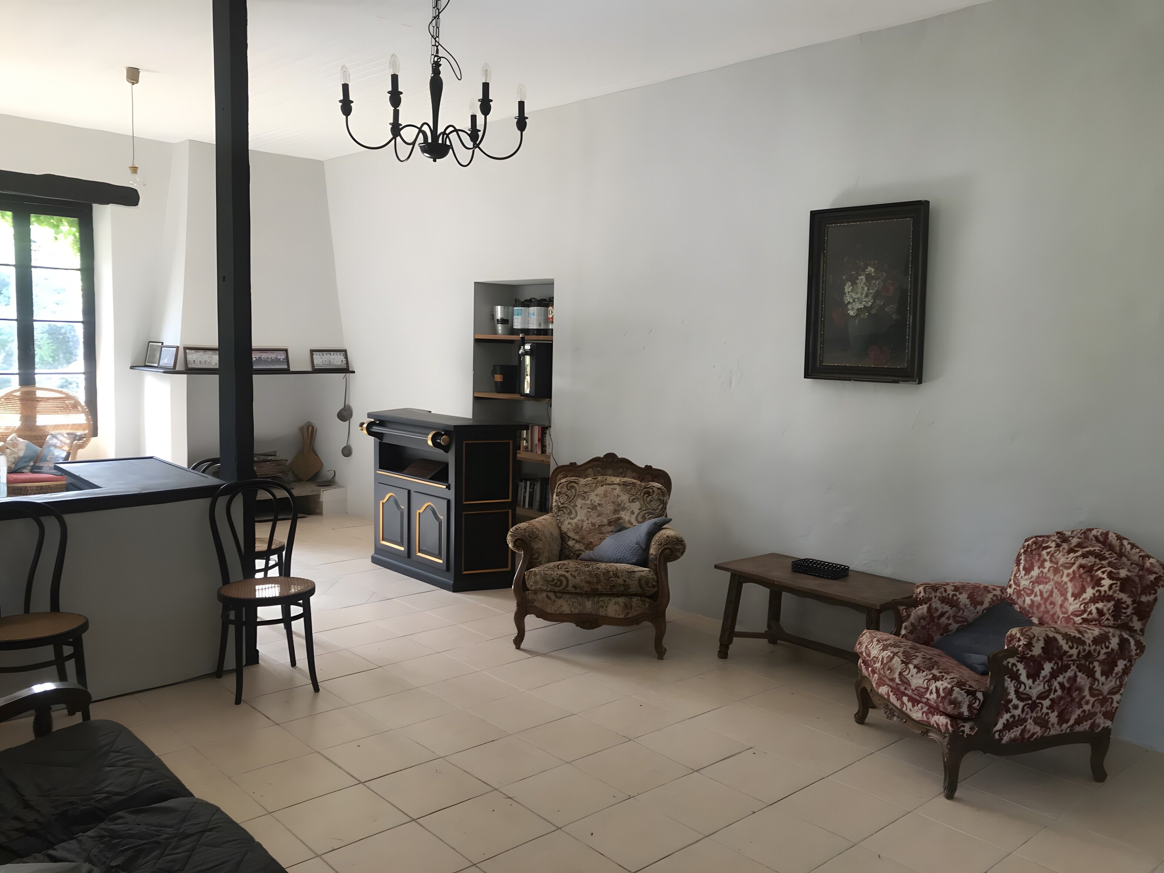 Sala de estar com sofá, poltronas, mesa pequena, lustre, lareira e área de cozinha com bancada e prateleiras.