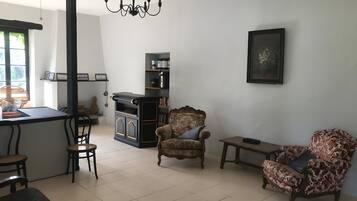 Sala de estar com sofá, poltronas, mesa pequena, lustre, lareira e área de cozinha com bancada e prateleiras.