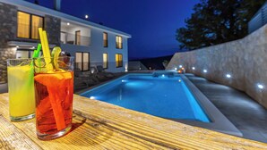 Blick auf einen Pool mit zwei Getränken, eine Steinsäule und ein modernes Haus im Hintergrund.