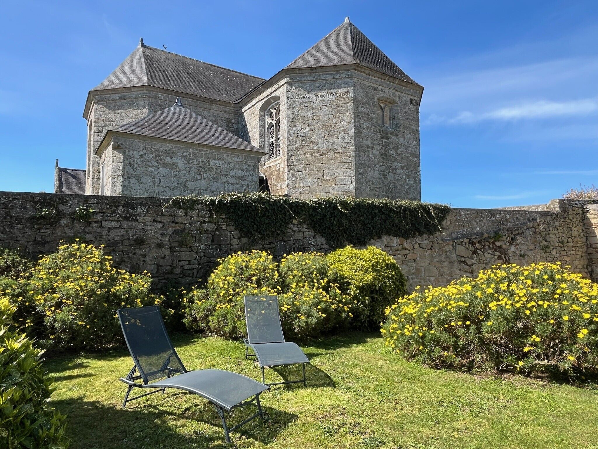 A stone building with a tower, a stone wall, and two metal chairs in a garden.