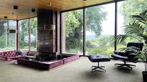 Een ruime woonkamer met een open haard, grote ramen en uitzicht op groen.