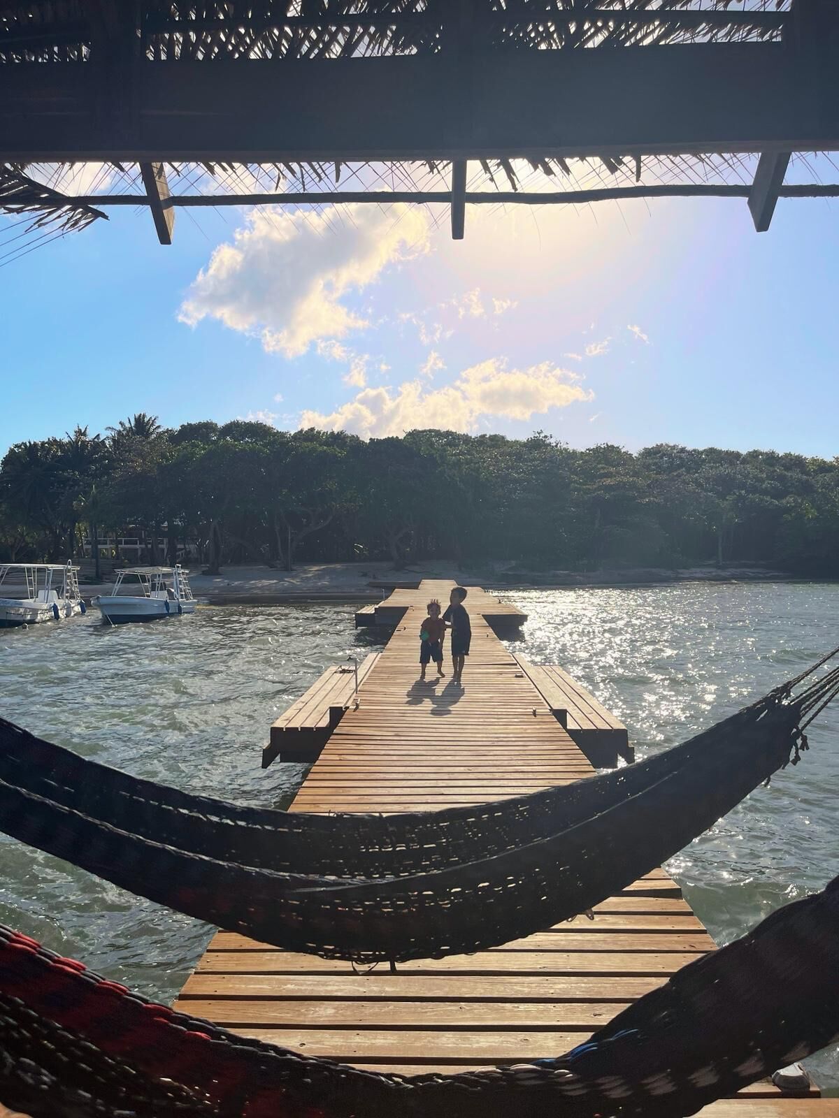 Een houten steiger die uitloopt in kalm water, met twee personen aan het uiteinde. Boten liggen aan de linkerzijde afgemeerd, en een rieten dakconstructie is zichtbaar.
