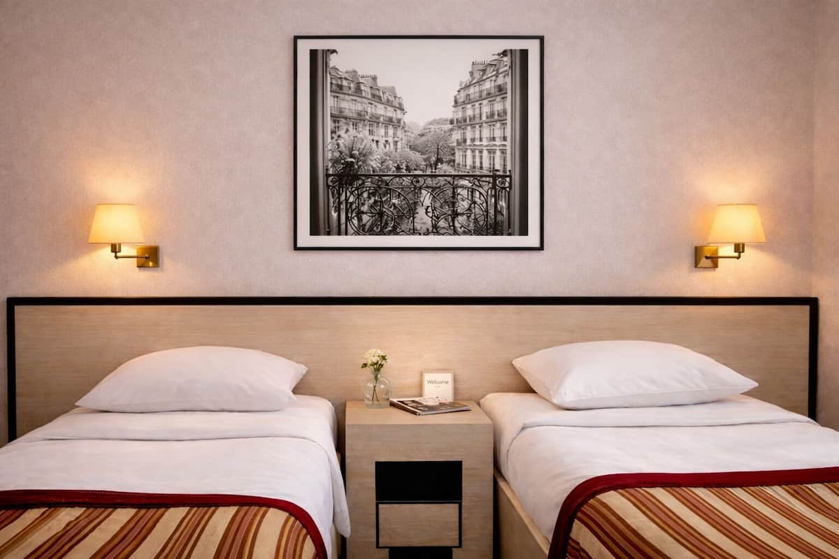 A hotel room with two beds, a framed cityscape picture, and two wall-mounted lamps.