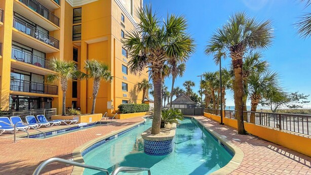 A hotel pool area with a clear blue pool, sun loungers, palm trees, and a yellow building facade. - St. John's Inn (Myrtle Beach)
