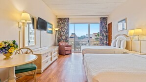A hotel room with a large bed, a desk with a chair, a small table with a vase of flowers, a television mounted on the wall, and a balcony with a view of buildings. - St. John's Inn (Myrtle Beach)