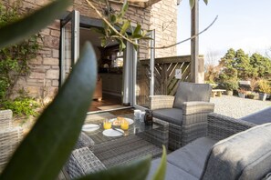 Un área de estar al aire libre con mobiliario de mimbre, una mesa de vidrio y vistas a la cocina a través de puertas de cristal.