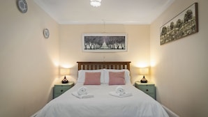 Een keurig opgemaakt bed met witte lakens, twee roze kussens en twee gevouwen handdoeken. Twee nachtkastjes met lampen, een ingelijst foto aan de muur en een plafondlamp.