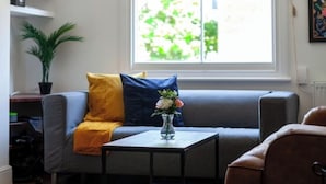 Una sala de estar con un sofá gris, una mesita con un jarrón de flores, una planta en maceta y una ventana con vistas al verdor.