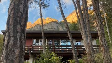 Eine Holzhütte mit einer Holzterrasse und großen Fenstern, umgeben von Bäumen und Bergen im Hintergrund.