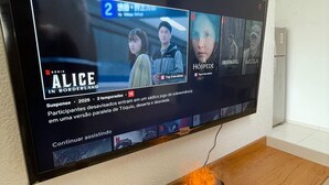 A television mounted on a wall with a wooden shelf below displaying a small black device emitting light.