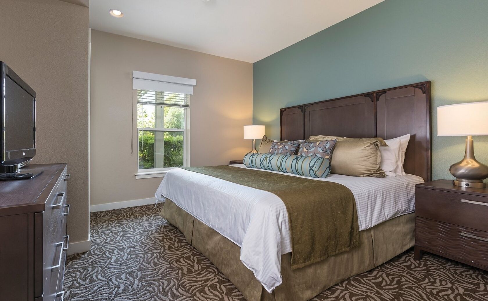 A bedroom with a bed, a TV, a window, and a patterned carpet.