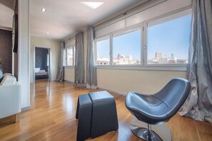 Ein geräumiges Zimmer mit einem modernen Sessel, einem kleinen Hocker und einem großen Fenster mit Blick auf die Stadt.