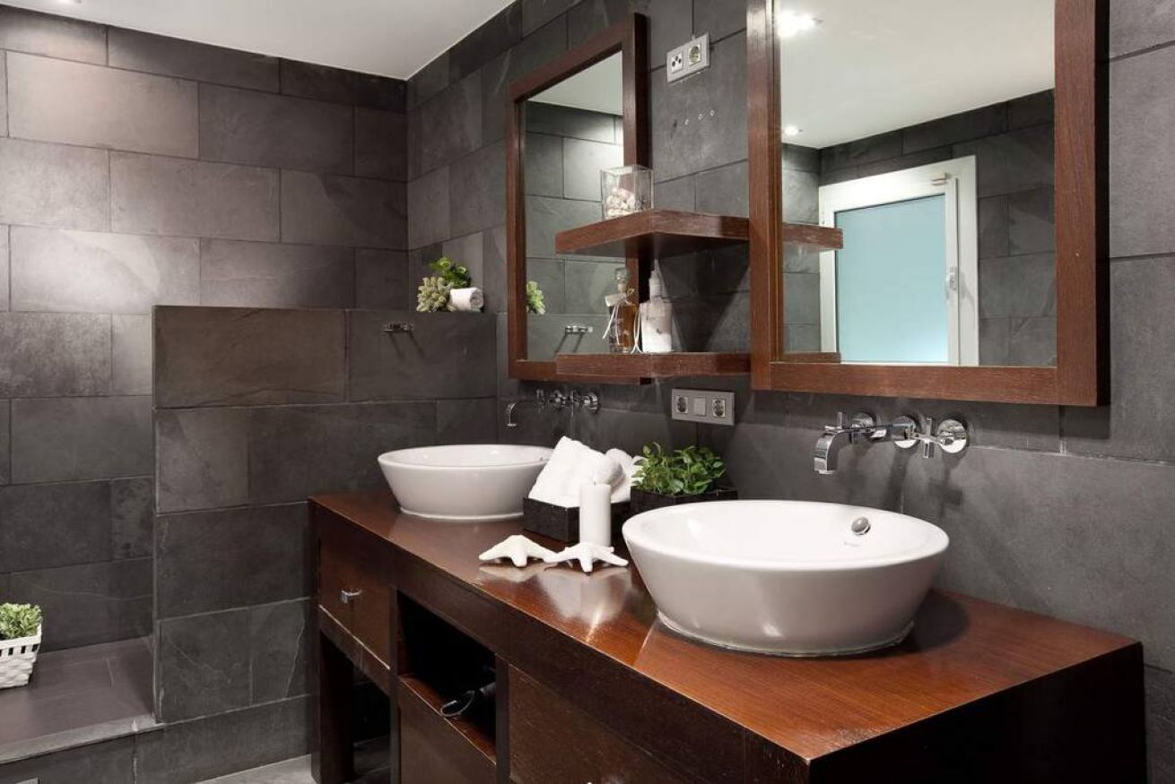 Un bagno con due lavandini, piastrelle in pietra scura, un mobile contenitore in legno e uno specchio.