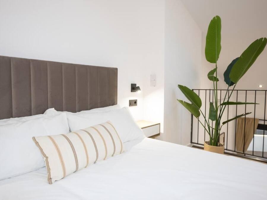 Una cama bien hecha con sábanas blancas y almohadas a rayas, un cabecero tapizado en gris y una planta en maceta sobre una mesita de noche.