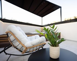A wicker lounge chair with white cushions on a black round table, a potted plant, and a modern outdoor seating area with a dark roof structure. - Dorian Suites (Monforte de Lemos)