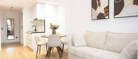 A modern living room with a sofa, a dining area with chairs, a kitchen, and abstract wall art.