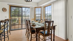 Un coin repas avec une table en bois, des chaises et un lustre. De grandes fenêtres, équipées de stores, offrent une vue sur l’extérieur.