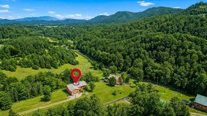 Un paisaje rural con una casa señalada por un puntero rojo, rodeada de un bosque denso y colinas ondulantes.