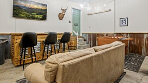 Une pièce chaleureuse avec un canapé, trois tabourets de bar noirs, un comptoir en bois, une tête de cerf fixée au mur, une porte verte, un tableau encadré et une table de baby-foot.
