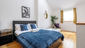 Un dormitorio con cama, mesita de noche, lámpara, ventana y una obra de arte.