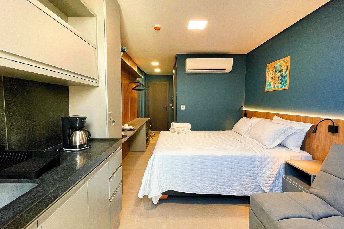 Una habitación de hotel compacta con kitchenette, una cama con ropa blanca, un pequeño rincón de descanso y un cuadro en la pared.