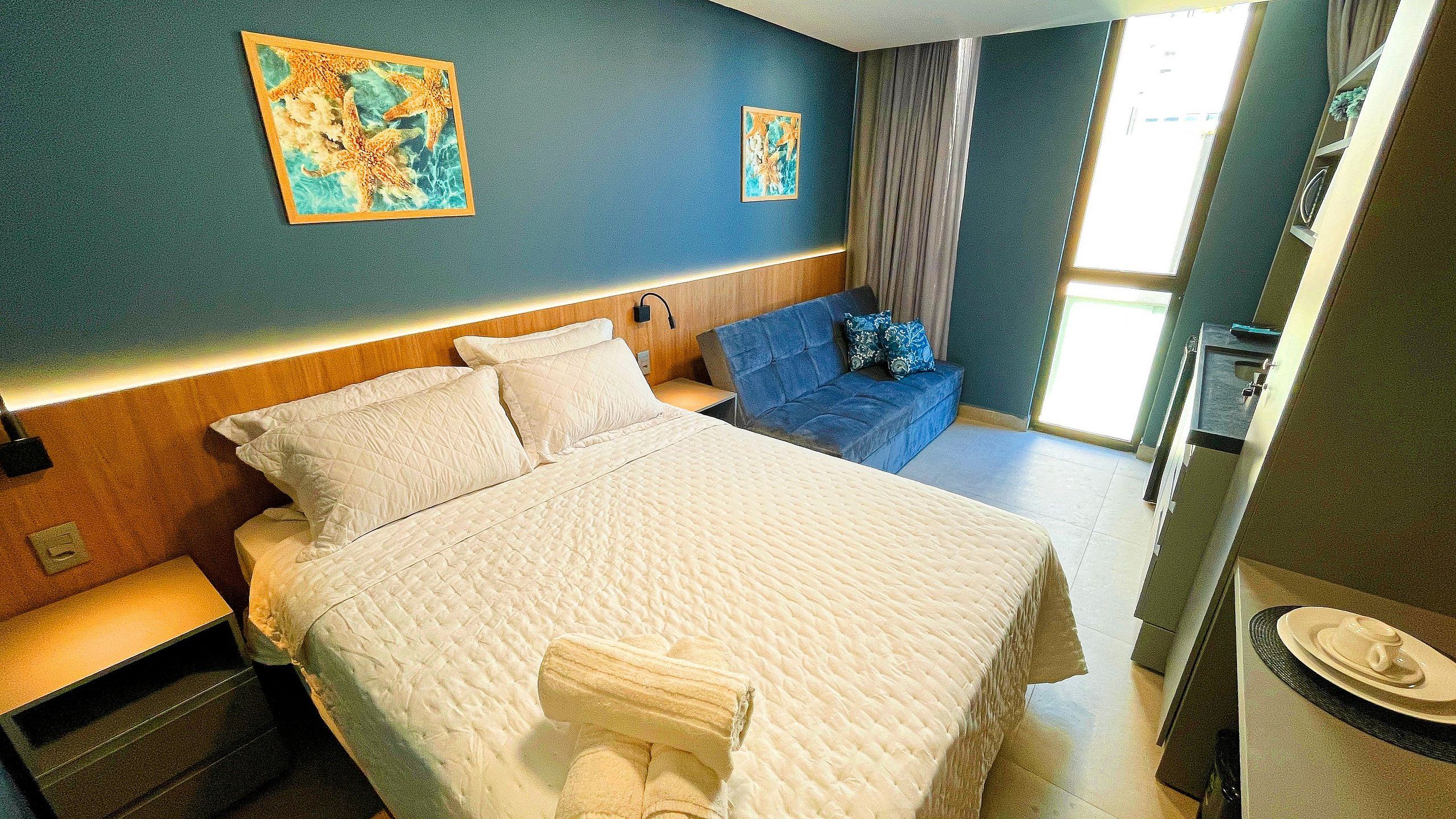 Een hotelkamer met een bed, een blauwe bank, een nachtkastje, een wandlamp en kunstwerken.