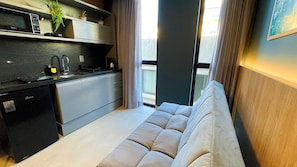 Una cocina moderna con microondas integrado, cafetera y un pequeño lavamanos. En la sala de estar hay un sofá gris con vistas al exterior a través de un ventanal amplio.