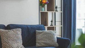En blå soffa med mönstrade kuddar, en vit hylla med dekorativa föremål och en bokhylla med färgglada blommor.