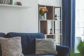 A blue sofa with patterned cushions, a white shelf with decorative items, and a bookshelf with colorful flowers. - Stylish Deluxe Two-Bedroom Apartment in Vibrant Vauxhall – Sleeps 6 (London)
