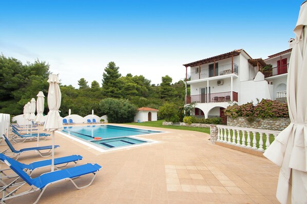 A pool area with lounge chairs and umbrellas, a swimming pool, and a building with balconies. - VILLA ALISSIA (Skiathos)