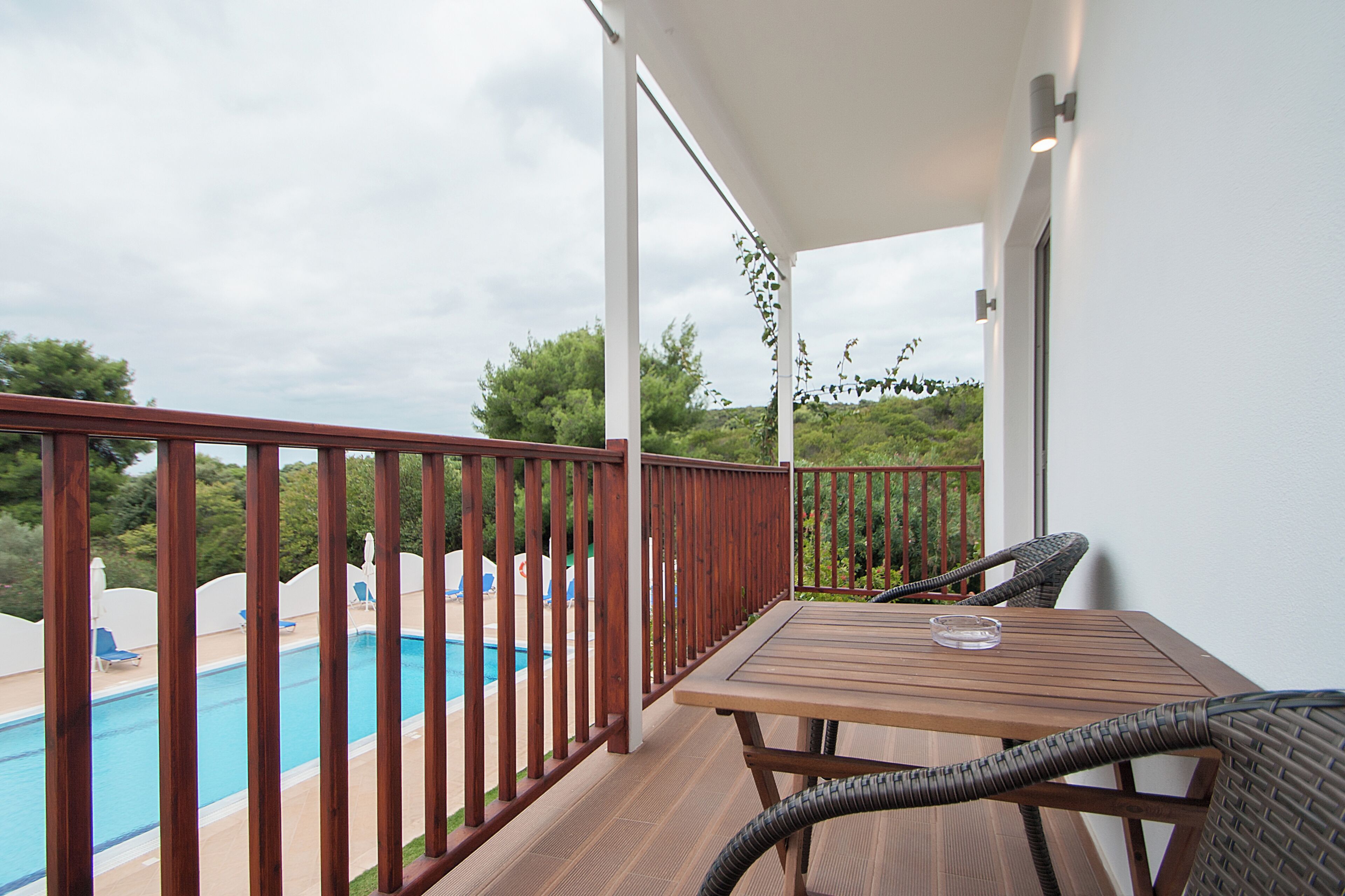 A balcony with a wooden table and wicker chairs, overlooking a swimming pool and greenery.