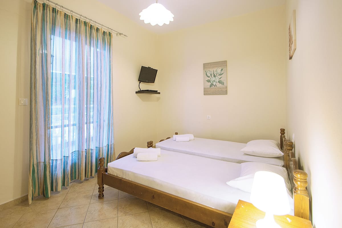 A room with two wooden beds, a television on a stand, a window with curtains, and a wall-mounted picture.