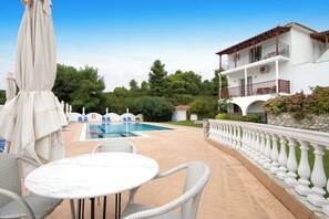 A poolside area with a white table and wicker chairs, a pool, and a building with a balcony. - VILLA ALISSIA (Skiathos)
