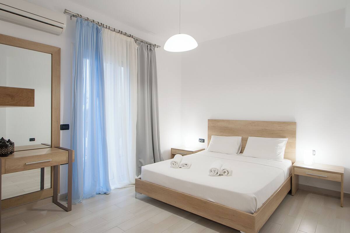 A modern hotel room with a wooden bed, a bedside table, a nightstand, a mirror, and a window with sheer curtains.
