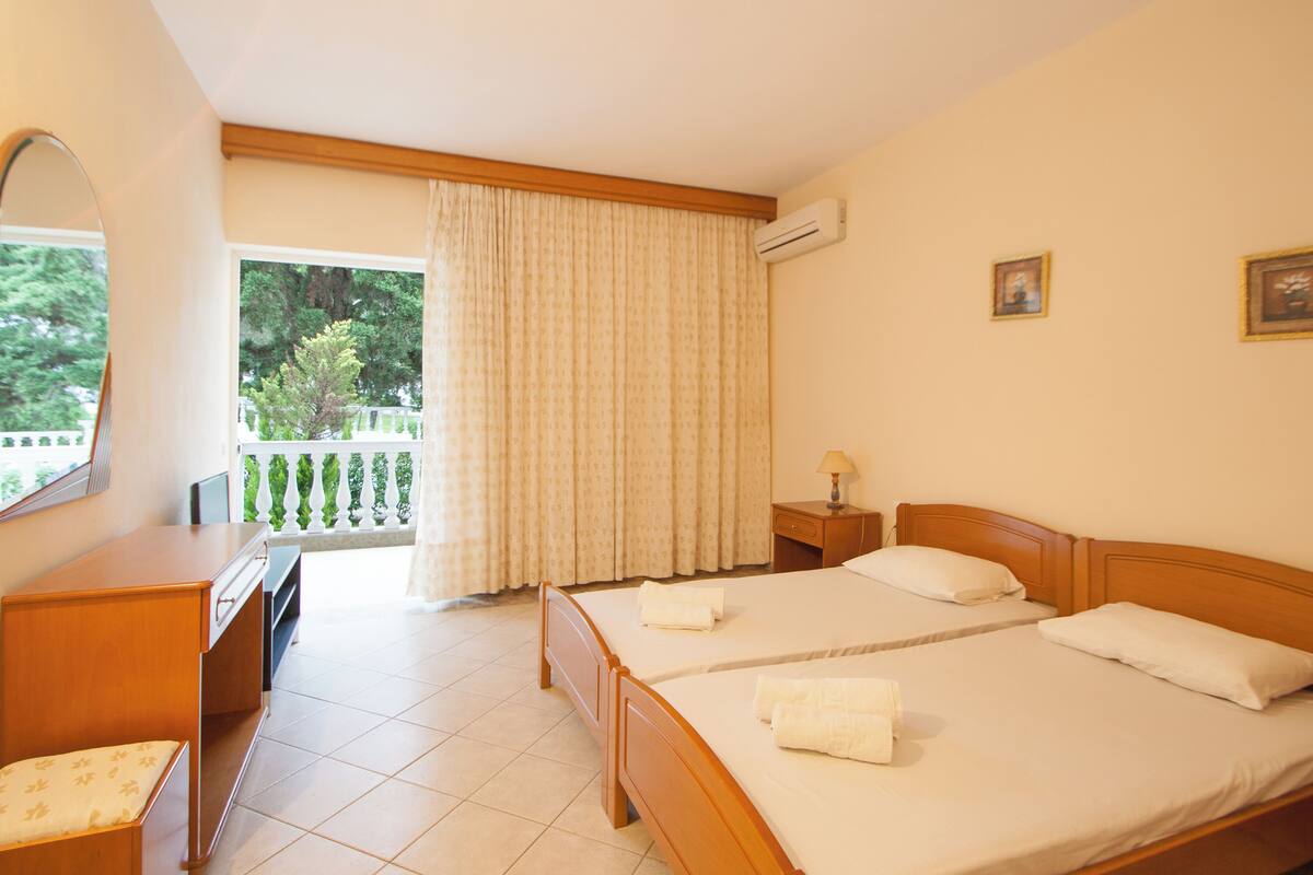 A hotel room with two beds, a desk, a chair, a window with curtains, and an air conditioning unit.