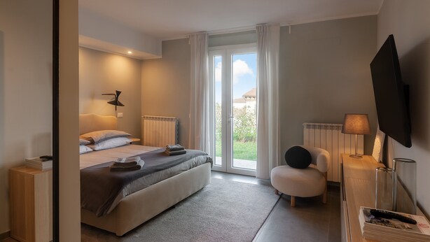 A modern bedroom with a bed, bedside tables, a chair, a TV, and a view of the outdoors. - Blueyes House Tuscany (Montespertoli)