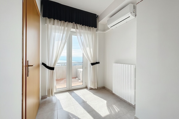 Una stanza con una porta-finestra scorrevole che dà su un balcone, un termosifone bianco e un condizionatore d'aria montato a parete.