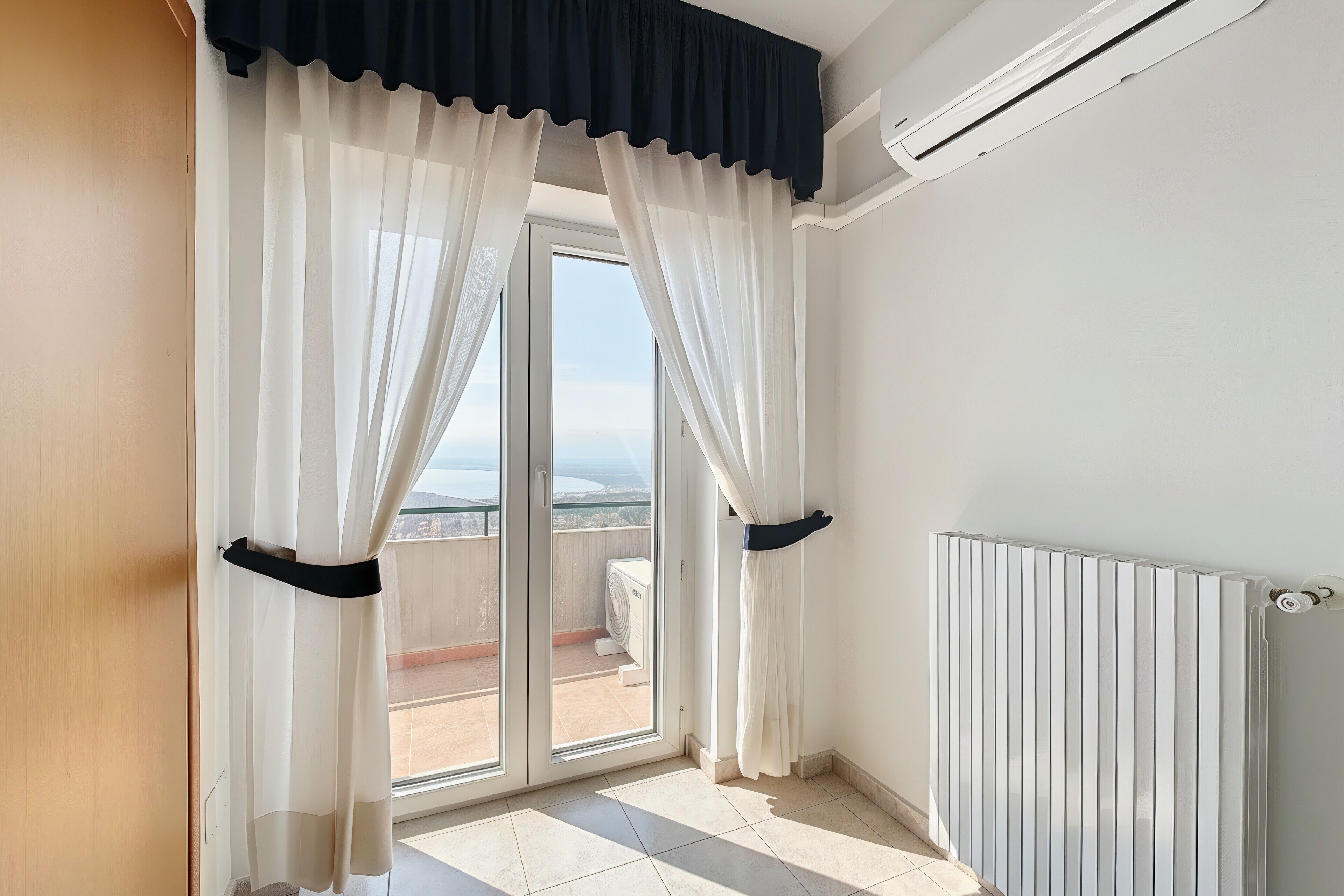 A room with a sliding glass door leading to a balcony, a radiator, and a view of the sky.