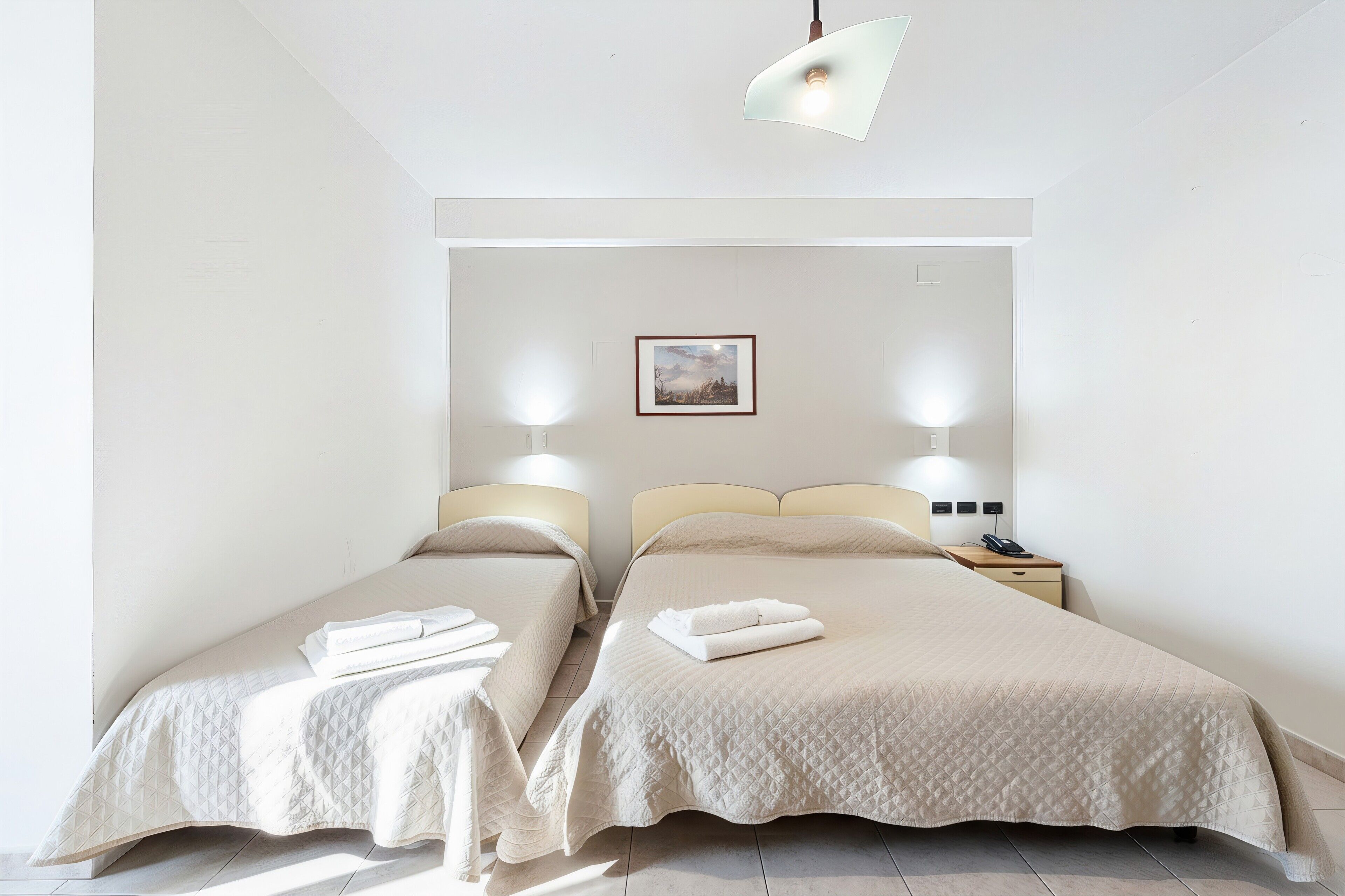 Une chambre d’hôtel avec deux lits, une table de chevet avec une lampe, un tableau encadré au mur et un luminaire au plafond.