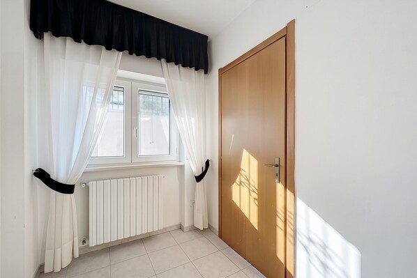 Quarto com janela, cortinas brancas, radiador e porta de madeira.