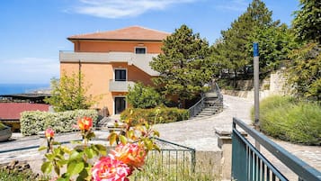 A multi-story building with a red-tiled roof, a staircase leading to a balcony, and a view of the sea.