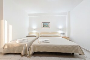 Ein Hotelzimmer mit zwei Betten, einem Nachttisch, einem Tischchen neben dem Bett, einem gerahmten Bild an der Wand und einem Fenster, das für natürliches Licht sorgt.