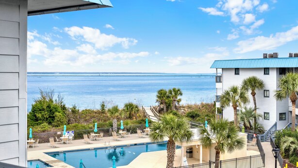 A pool area with lounge chairs and umbrellas, a view of the ocean, and a building with a blue roof. - Fully equipped, 1 bedroom, 1 bath Bay front condo on Tybee Island with a pool (Tybee Island)