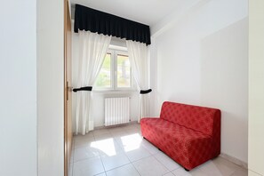 Una habitación con sofá rojo, cortinas blancas, una ventana y un radiador.