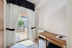 Et værelse med et træskrivebord, en radiator, et vindue med hvide gardiner og udsigt til det fri.