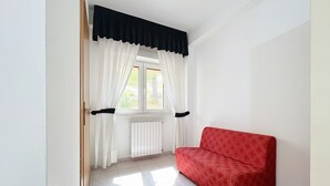 Une pièce avec un canapé rouge, des rideaux blancs, une fenêtre et un radiateur.