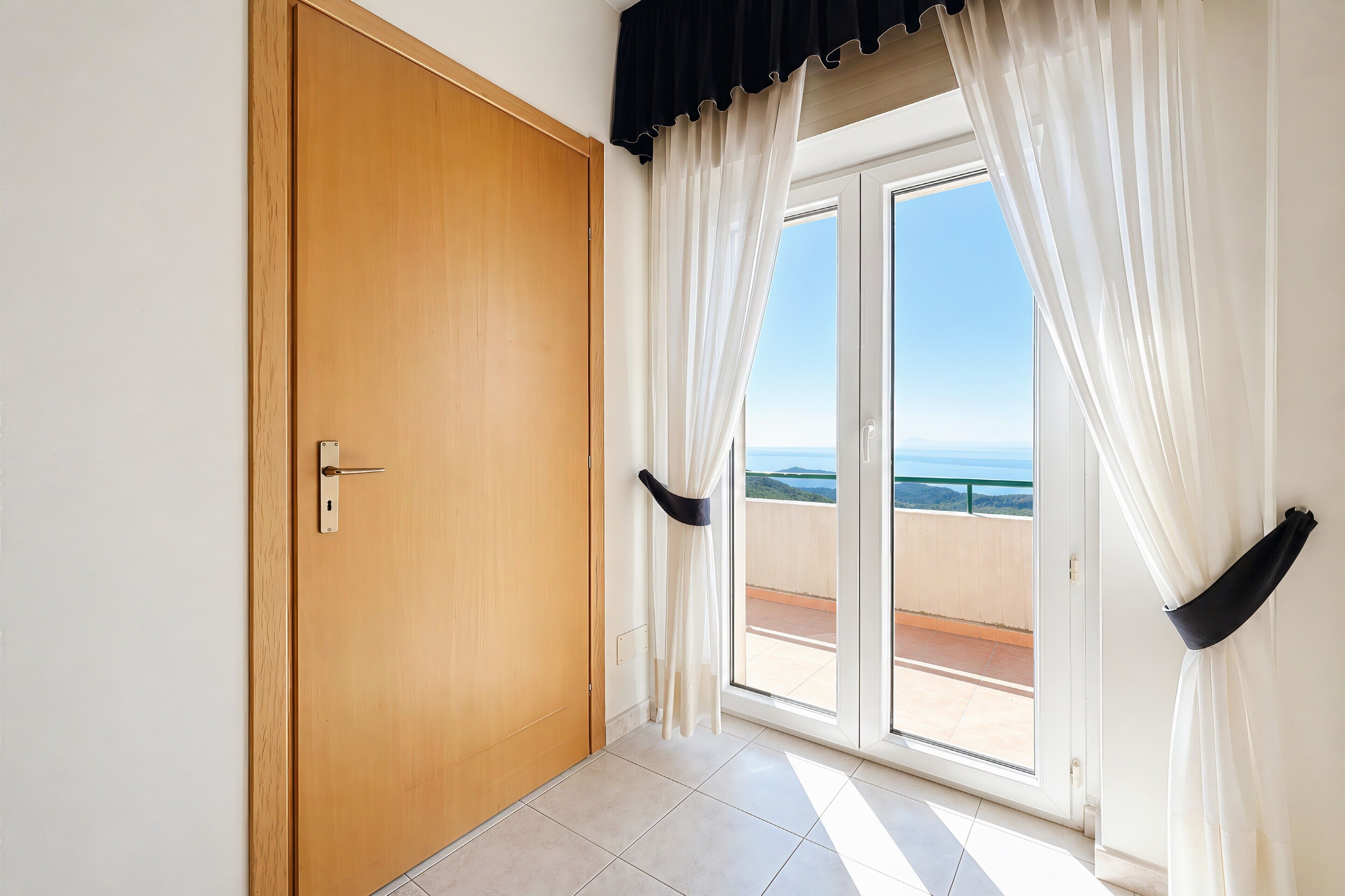 A room with a wooden door, a sliding glass door to a balcony, and sheer curtains.